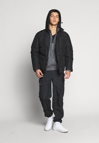 Black padded jacket with a hood, featuring a zipper and side pockets, paired with dark cargo pants and white sneakers. Smooth texture.