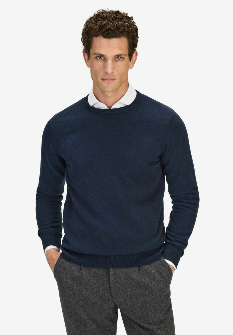 Marineblå stickad tröja med rund hals i mjukt tyg, med ribbade muddar och nederkant, bärs över en vit skjorta med krage.