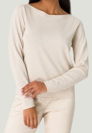 Femme portant un haut blanc texturé à manches longues avec de petits points en relief et un pantalon blanc, les bras croisés à la taille.