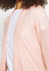 Světle růžový kardigan se zapínáním vpředu, vyrobený z měkkého materiálu a s dlouhými rukávy. Nosený přes bílé tričko, má uvolněný střih a jemný design.