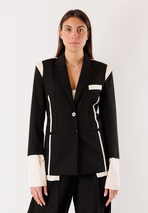Donna con blazer nero a righe bianche e polsini bianchi allungati, in piedi davanti a uno sfondo bianco uniforme.
