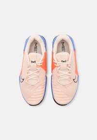 Zapatillas deportivas de malla en color melocotón con acentos en naranja y azul. Las características incluyen una parte superior texturizada, cordones blancos y un logo negro en el costado.