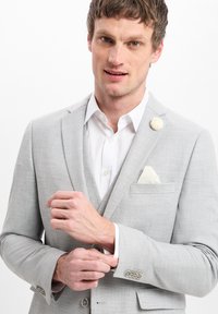Helle graue strukturierte Blazer mit Revers, weißem Taschentuch und einem Stoffblumen-Anstecker. Sichtbare Knopfriegeldetails. Darunter ein weißes Hemd.