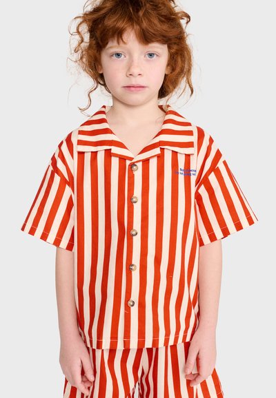 Enfant aux cheveux roux portant une chemise à manches courtes boutonnée et un short assorti avec des rayures verticales rouges et blanches sur un fond uni.