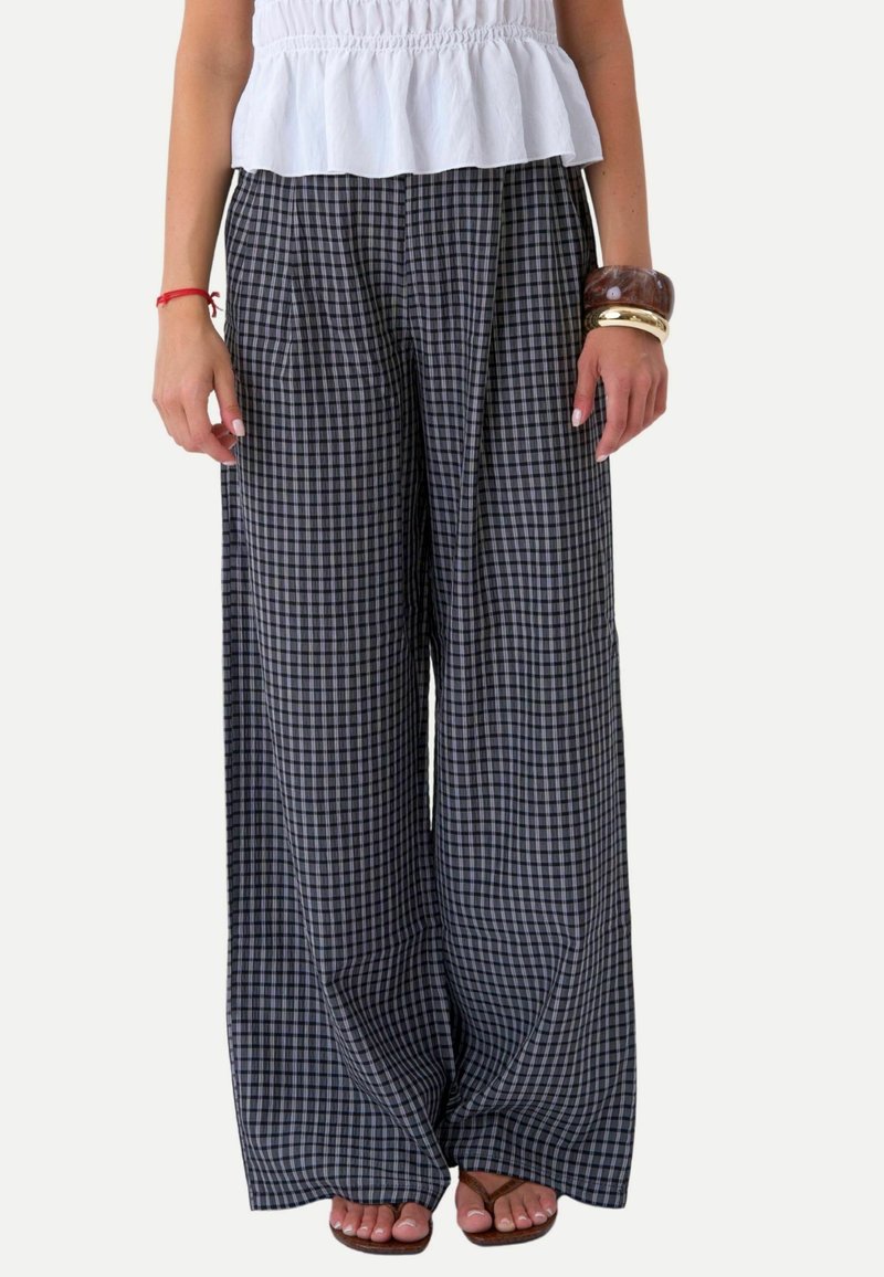 Femme portant un pantalon large à carreaux noirs et blancs, un top blanc sans manches, des bracelets et des sandales marron, debout devant un fond uni.
