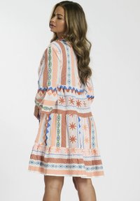 Donna con i capelli lunghi e mossi, che indossa un vestito multicolore a stampa lungo fino al ginocchio, in piedi e che guarda oltre la spalla su uno sfondo bianco.