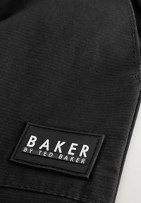 Schwarzer Stoff mit einem rechteckigen schwarzen Etikett, auf dem in weißen Buchstaben "BAKER BY TED BAKER" genäht ist.