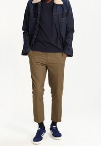 Person iført khaki bukser, marineblå sneakers med hvide striber, marineblå skjorte og mørk jeansjakke med shearling krave, hænderne i lommerne.
