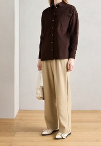 Camicia marrone con bottoni e tasca sul petto, abbinata a pantaloni beige a gamba larga e scarpe bianche slip-on. Lo sfondo è neutro.