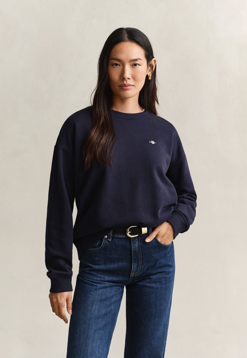 Kvinde med langt mørkt hår iført navy sweatshirt og blå jeans med sort bælte, står mod en ensfarvet lys baggrund.
