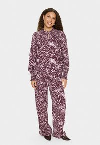 Donna in piedi che sorride, indossa una blusa ampia a maniche lunghe con un motivo viola e rosa abbinato e pantaloni larghi, completando l'outfit con scarpe nere a punta.