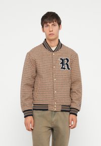 Brun houndstooth bomberjakke med marineblå ribbestrikket krage og mansjetter, med et fremtredende "R"-logo og trykknap-lukking.