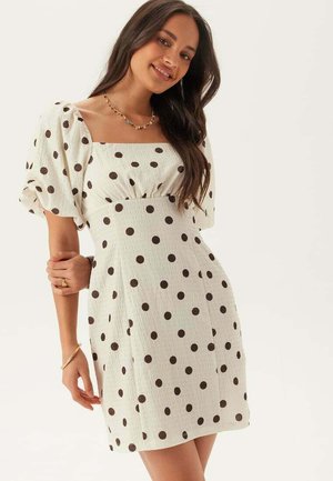 Giovane donna con capelli lunghi e scuri che indossa un vestito bianco con pois neri e maniche a sbuffo, sorridente con un braccio piegato.