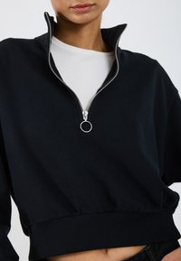 Sweat à zip noir fabriqué en tissu doux avec un col montant, présentant une fermeture éclair en argent et un ourlet côtelé. Design simple et décontracté.