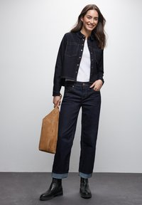Giacca di jeans scura con bottoni in rame, t-shirt bianca, jeans scuri con risvolti, e stivaletti neri. Tiene una borsa a tracolla in suede beige.