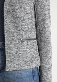Šedá texturovaná bunda v cropped designu se stříbrným zipovým kapsičkem. Spárována s modrými džíny, které mají přiléhavý střih.
