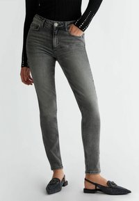 Vaqueros ajustados grises con un diseño desgastado, cinco bolsillos y cierre de botón metálico. Llevados con zapatos negros de textura sin cordones.