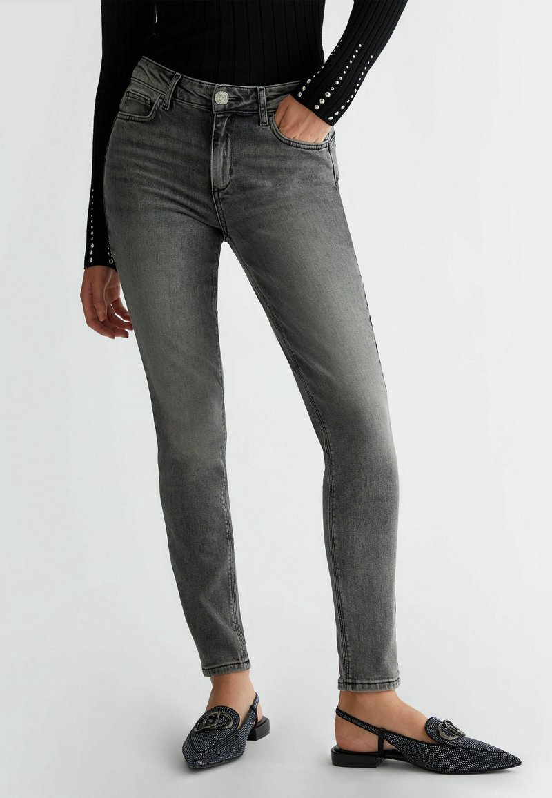 Vaqueros ajustados grises con un diseño desgastado, cinco bolsillos y cierre de botón metálico. Llevados con zapatos negros de textura sin cordones.