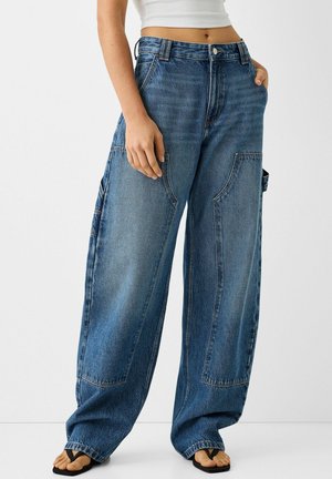 Osoba nosząca szerokie, niebieskie dżinsy z dużymi przednimi i bocznymi kieszeniami, w połączeniu z białym krótko skrojonym topem i czarnymi klapkami.