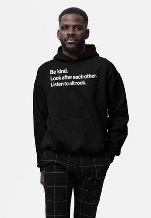 Mand iført en sort hættetrøje med hvid tekst "Vær venlig. Pas på hinanden. Lyt til alt rock." og sorte bukser med hvide gitterlinjer.