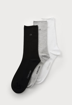 Tri osnovne Calvin Klein čarape s okruglim izrezom u crnoj, sivoj i bijeloj boji, svaka prikazuje "ck" logo blizu vrha i naziv brenda sa strane.