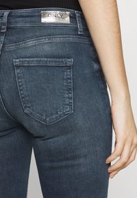 Vaqueros de mezclilla azul oscuro con una etiqueta de marca plateada en la cinturilla trasera y un único bolsillo trasero en forma de diamante. Textura suave, forma ajustada.