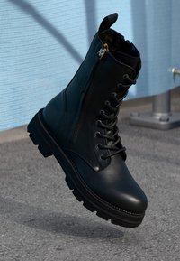 Stivale alla caviglia in pelle nera con lacci e chiusura con zip, caratterizzato da una finitura testurizzata e una suola in gomma spessa e scanalata.