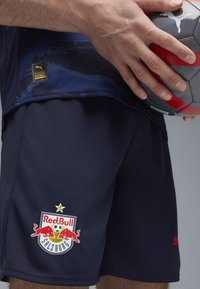Pantalones cortos deportivos en color azul marino, fabricados con un tejido transpirable, que cuentan con el logotipo de Red Bull Salzburg, una estrella y detalles en rojo. Una mano sostiene una pelota gris y roja.