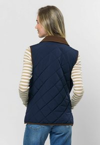 Námořnický modrý prošívaný vest s hnědým kordovým límcem, se hladkým povrchem a diamantovým vzorem, nošený přes pruhovanou košili s dlouhým rukávem.