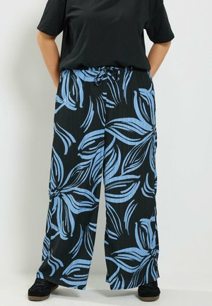 Persoon draagt losse zwarte broek met grote blauwe bloemenpatronen en zwarte schoenen, staand tegen een effen lichte achtergrond.