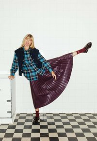 Chemise à carreaux bleue et verte, gilet foncé, jupe plissée violette brillante, chaussettes bordeaux avec des lettres blanches, et chaussures noires brillantes sur un sol à damier.