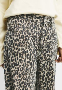Džins hlače z leopardjim vzorcem na svetlo bež podlagi, z temno rjavimi pikami, tradicionalnimi žepi in kovinskimi dodatki.