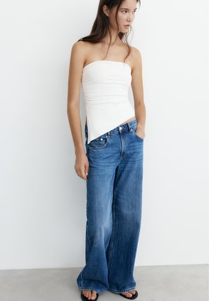 Jeune femme portant un top blanc asymétrique sans bretelles et un jean bleu à jambes larges, debout avec une main dans la poche contre un mur blanc.
