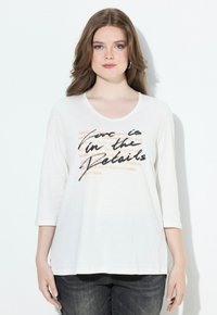 Donna con lungo capelli castani, indossa una camicia bianca a maniche tre quarti con testo nero e arancione, abbinata a jeans scuri, in piedi contro un muro bianco.