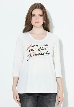 Vrouw met lang bruin haar draagt een witte shirt met driekwartmouwen met zwarte en oranje tekst, gecombineerd met donkere spijkerbroek, staande tegen een witte muur.