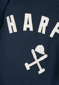 Sudadera azul marino con texto bordado en blanco que dice "HARPA" y un gráfico de un casco y palos cruzados. Textura suave.