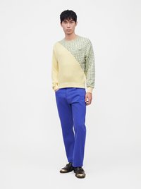 Homme portant un pull jaune et vert à motifs, un pantalon bleu et des chaussures noires avec des bouts dorés, debout avec une main dans la poche.