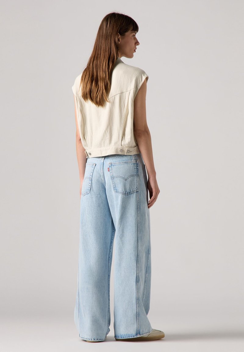 Débardeur en crème clair sans manches, design court, associé à un jean bleu clair ample avec deux poches arrière et une texture déchirée.