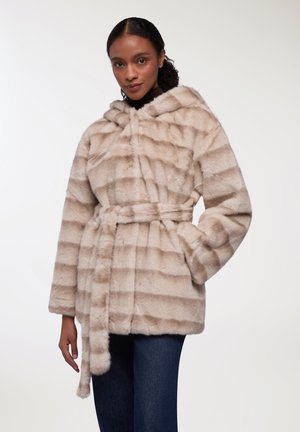 Cappotto in pelliccia sintetica beige con cappuccio, caratterizzato da strisce orizzontali e vita abbottonata. Tessuto morbido e maniche lunghe, indossato sopra dei jeans.
