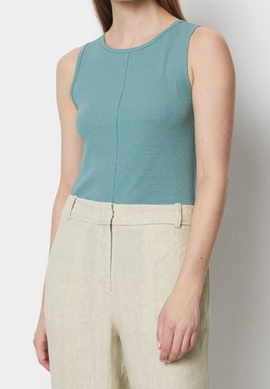 Mouwloos aansluitend teal topje gecombineerd met hooggetailleerde lichtbeige broek bij een staande persoon tegen een witte achtergrond.