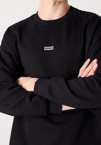 Persona vestida con una camisa de manga larga negra con el logo de "HUGO" en el pecho, con los brazos cruzados, visible desde el cuello hasta la mitad del torso contra un fondo liso.