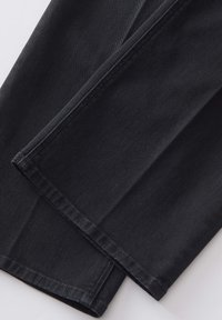 Gros plan sur des jambes de pantalon en denim noir avec des coutures visibles et des ourlets repliés sur un fond clair.