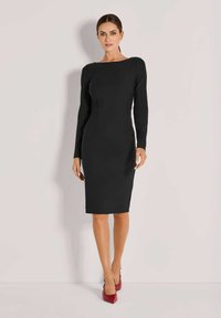 Frau in einem enganliegenden schwarzen Langarmkleid bis zum Knie und roten High Heels vor einem schlichten hellen Hintergrund.