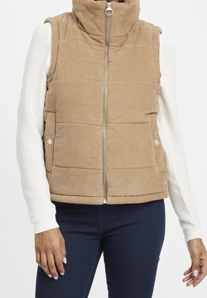 Gilet matelassé en velours côtelé beige avec fermeture éclair devant et col montant, porté sur un top blanc à manches longues côtelé et un pantalon bleu foncé.