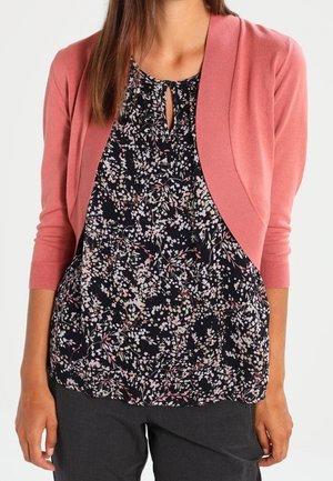 Kvinde iført en sort blomstret bluse med nøglehul-halsudskæring og en kort pink cardigan med trekvartlange ærmer.