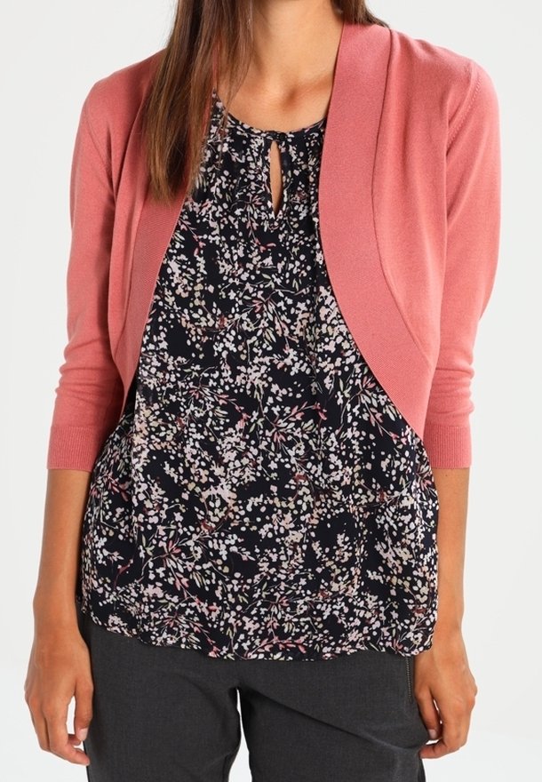 Femme portant un chemisier noir à motif floral avec une encolure en forme de trou de serrure et un cardigan court rose à manches trois-quarts.