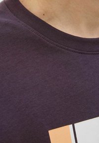 Camiseta de algodón morado oscuro con escote redondo. Presenta un estampado geométrico en colores claros, con costuras visibles a lo largo del collar.