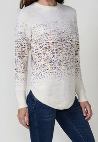 Femme portant un pull en tricot blanc avec un motif tacheté multicolore et un jean bleu foncé, visible du cou aux hanches.