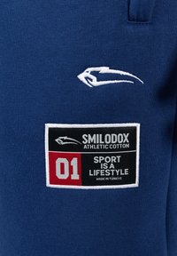 Blaue sportliche Baumwollkleidung mit einem weißen Logo und einem rechteckigen Patch mit schwarzem, rotem und weißem Text, der die Marke und den Slogan beschreibt.