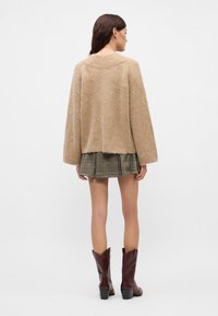 Mulher vestindo um suéter bege felpudo, uma saia plissada xadrez e botas de salto castanhas, de pé de costas contra um fundo simples.